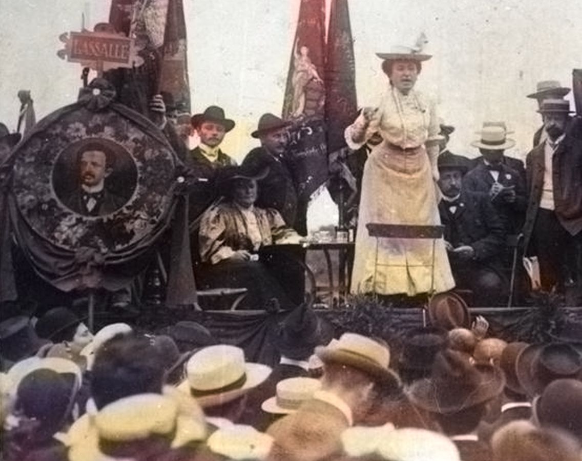 Foto von Rosa Luxemburg wie sie auf einer Bühne eine Rede vor einer Gruppe von Menschen hält, nachkoloriert mit KI